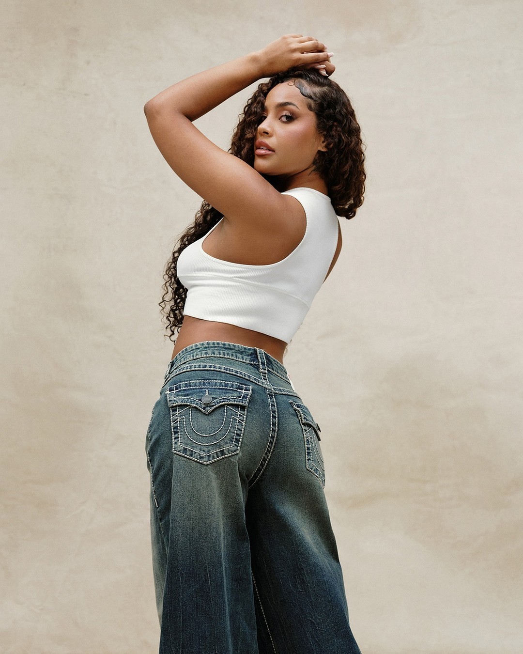 Knees-up portrait of a female model in jeans and a tank top with her hands abover her head and her body twisting towards the camera.